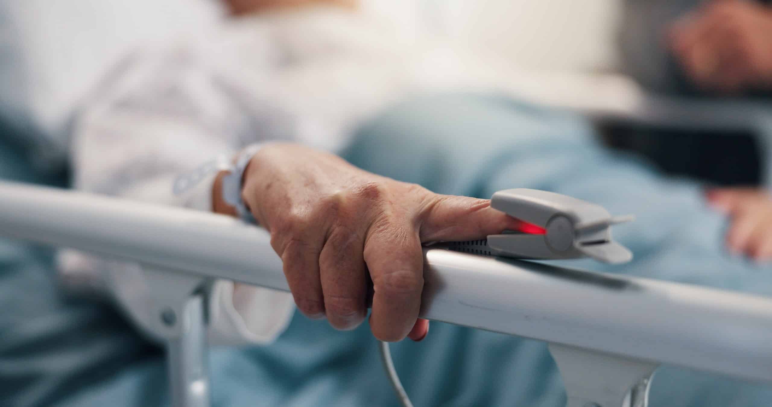 Patient hand lying on a hospital bed rail with a pulse oximeter attached.