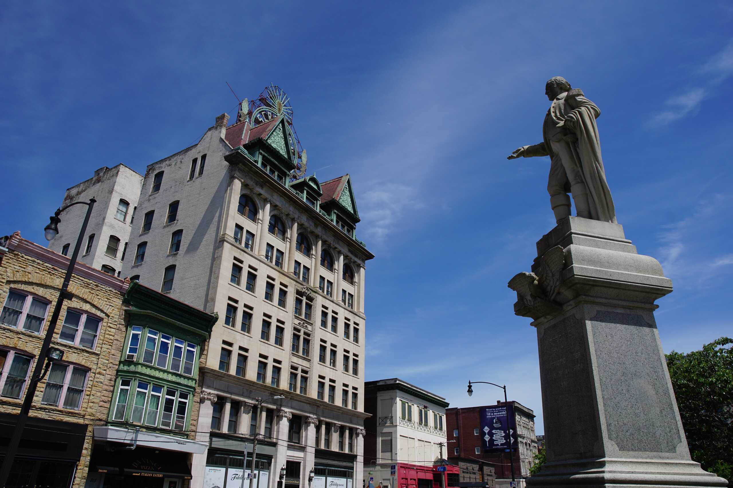 A stone historical monument stands in a town square under a bright blue sky with old architecture in the background, representing the area served by Scranton Burn Injury Lawyers.