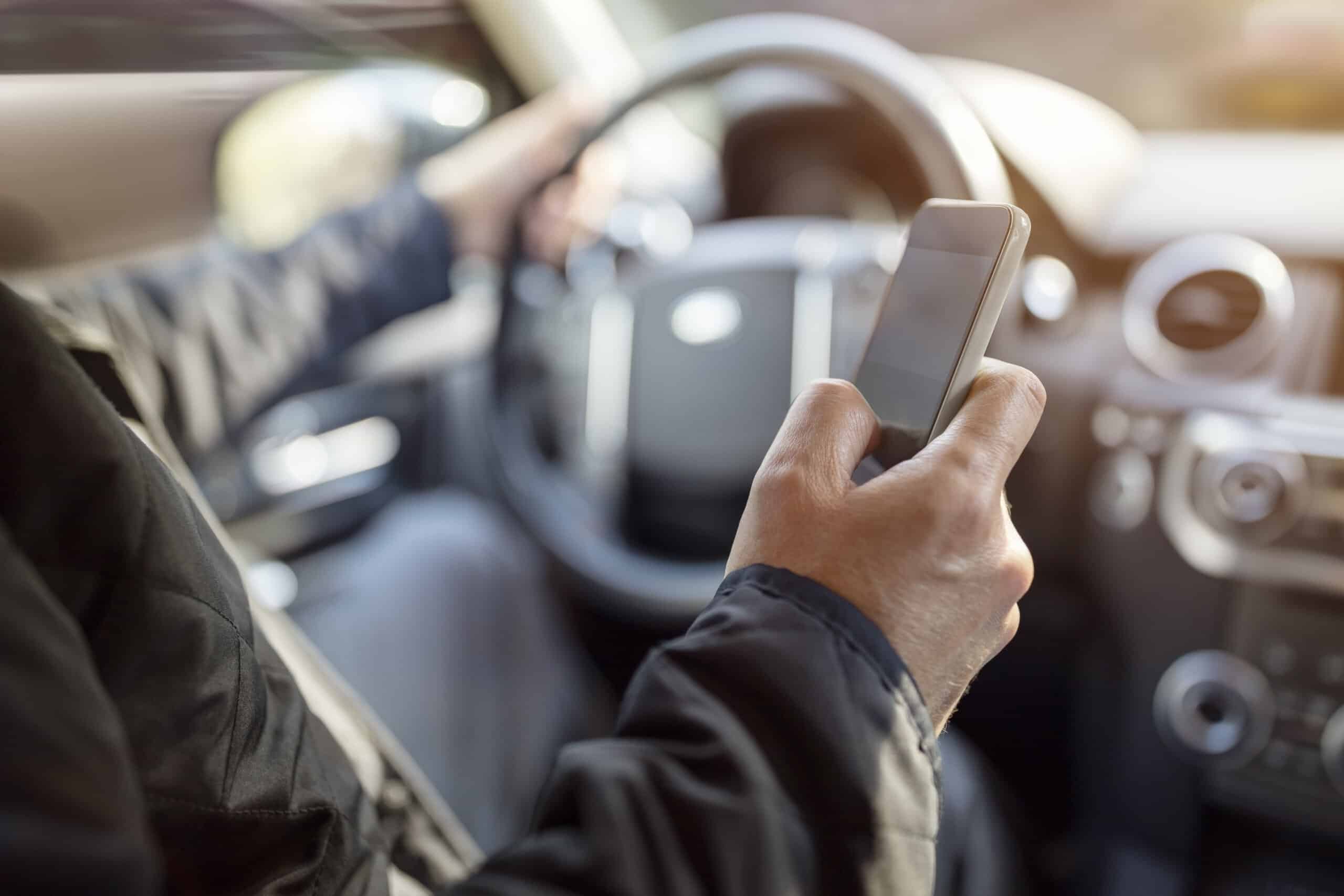 A close-up of a driver's hand holding and using a smartphone while behind the steering wheel of a moving vehicle, illustrating a case handled by Scranton distracted driving accident lawyers.