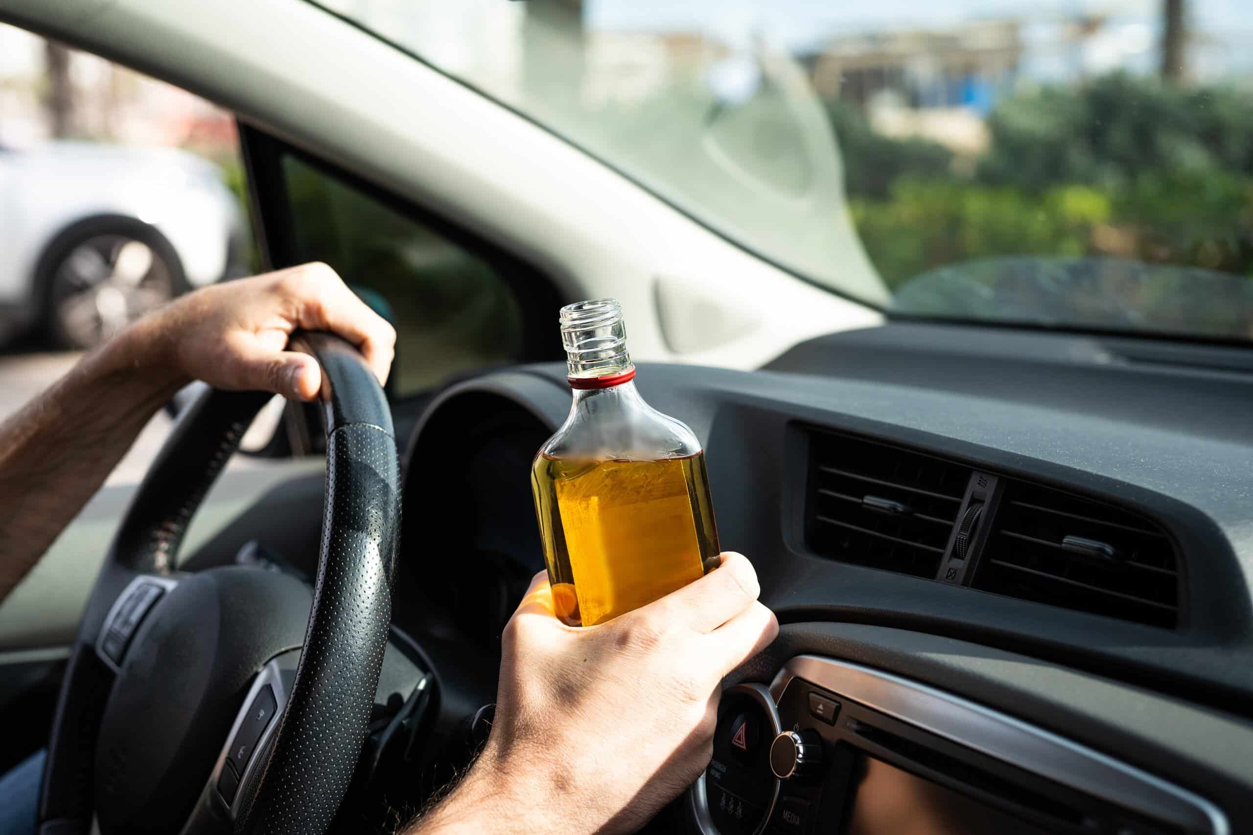 A driver holding a steering wheel with one hand and an open bottle of liquor with the other inside a vehicle, illustrating a case handled by Scranton Drunk Driving Accident Lawyers.