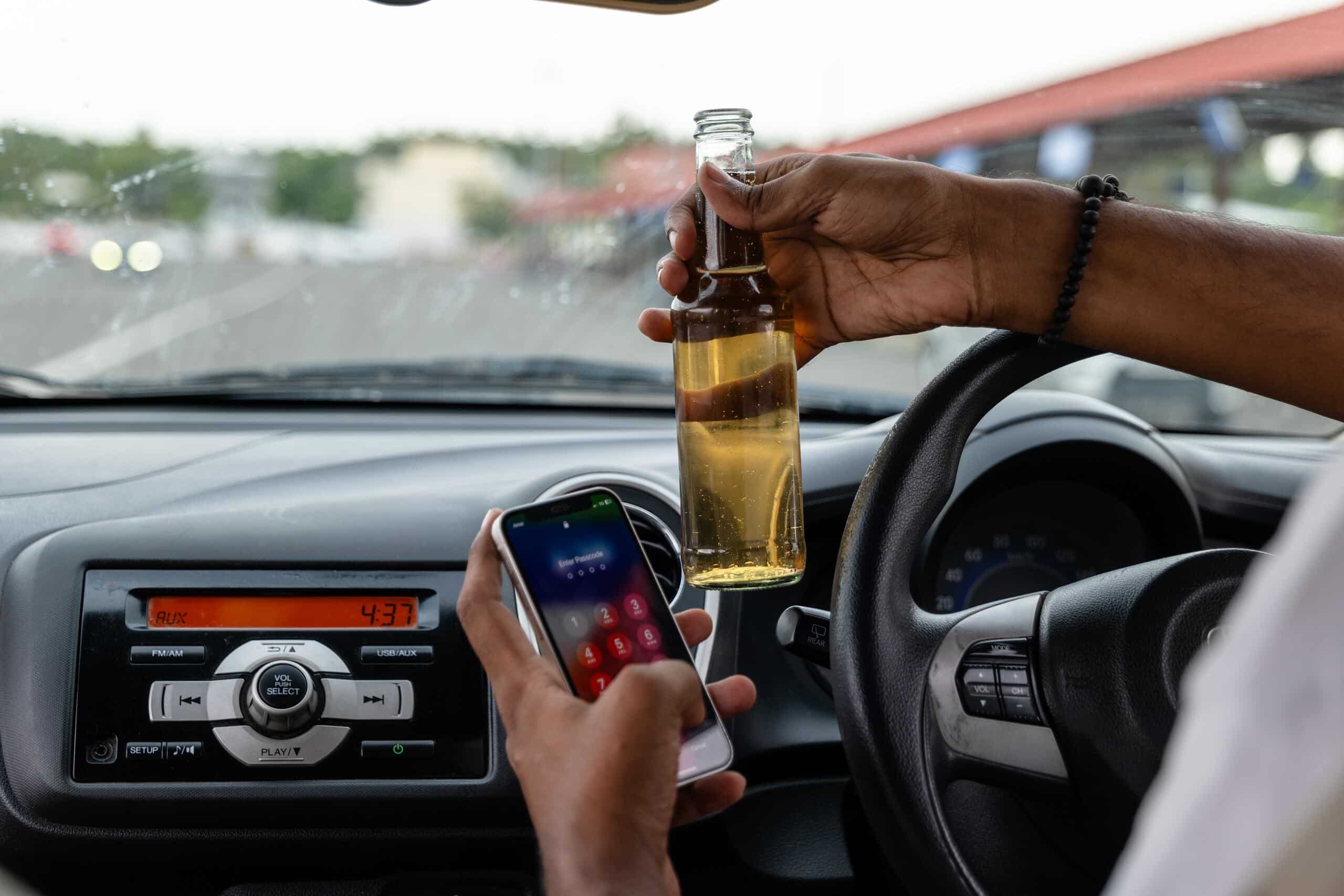 A driver holding a glass beer bottle by the steering wheel while using a smartphone with the other hand, highlighting reckless behaviors addressed by Scranton Drunk Driving Accident Lawyers.