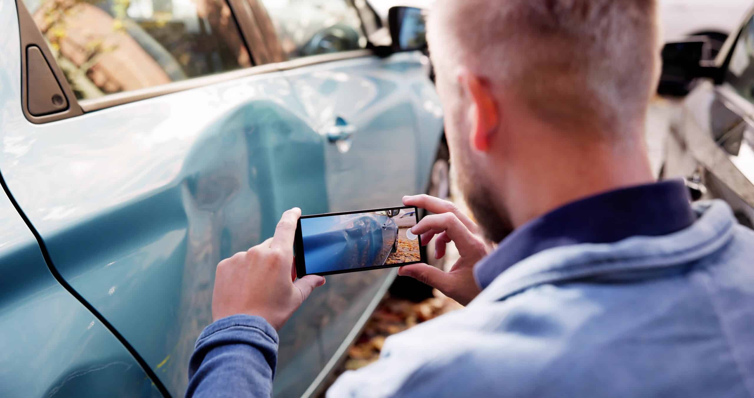 A man using a smartphone to document a large dent on the side of a blue car, collecting evidence for Scranton Uninsured Motorist Accident Lawyers.