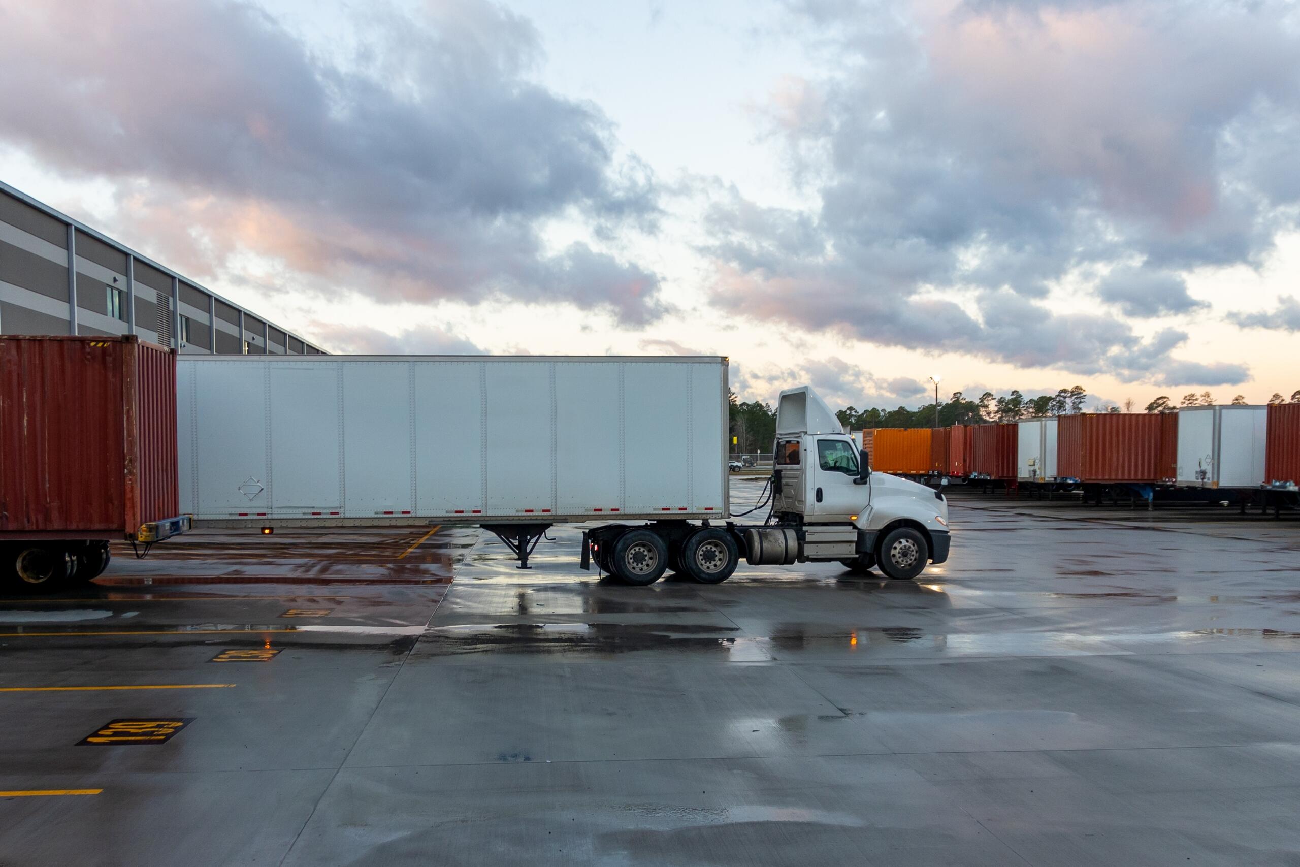White semi truck maneuvers in a large industrial shipping yard.